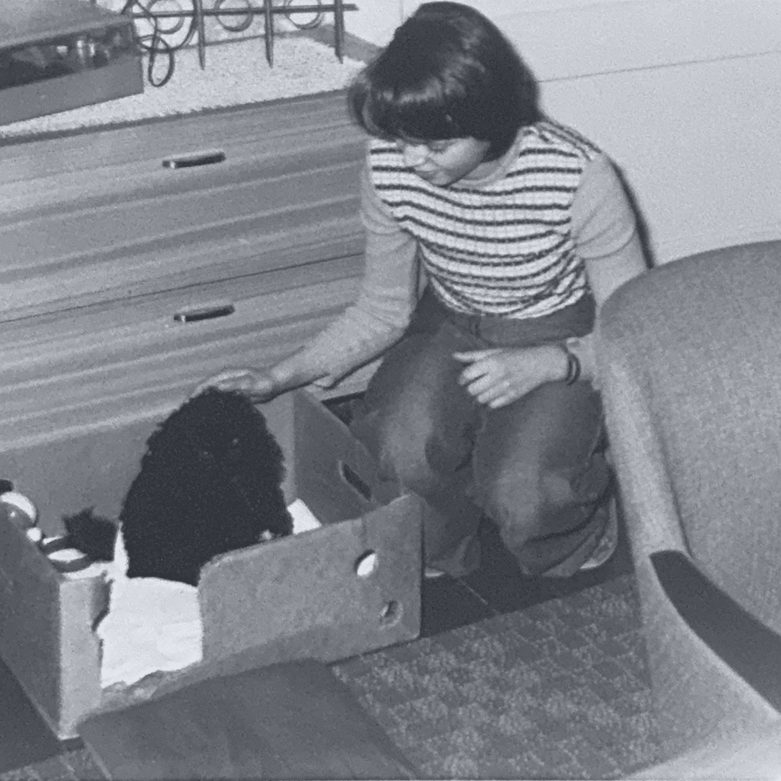 Ramona als junges Mädchen streichelt den in einer Kiste sitzenden schwarzen Pudelwelpen