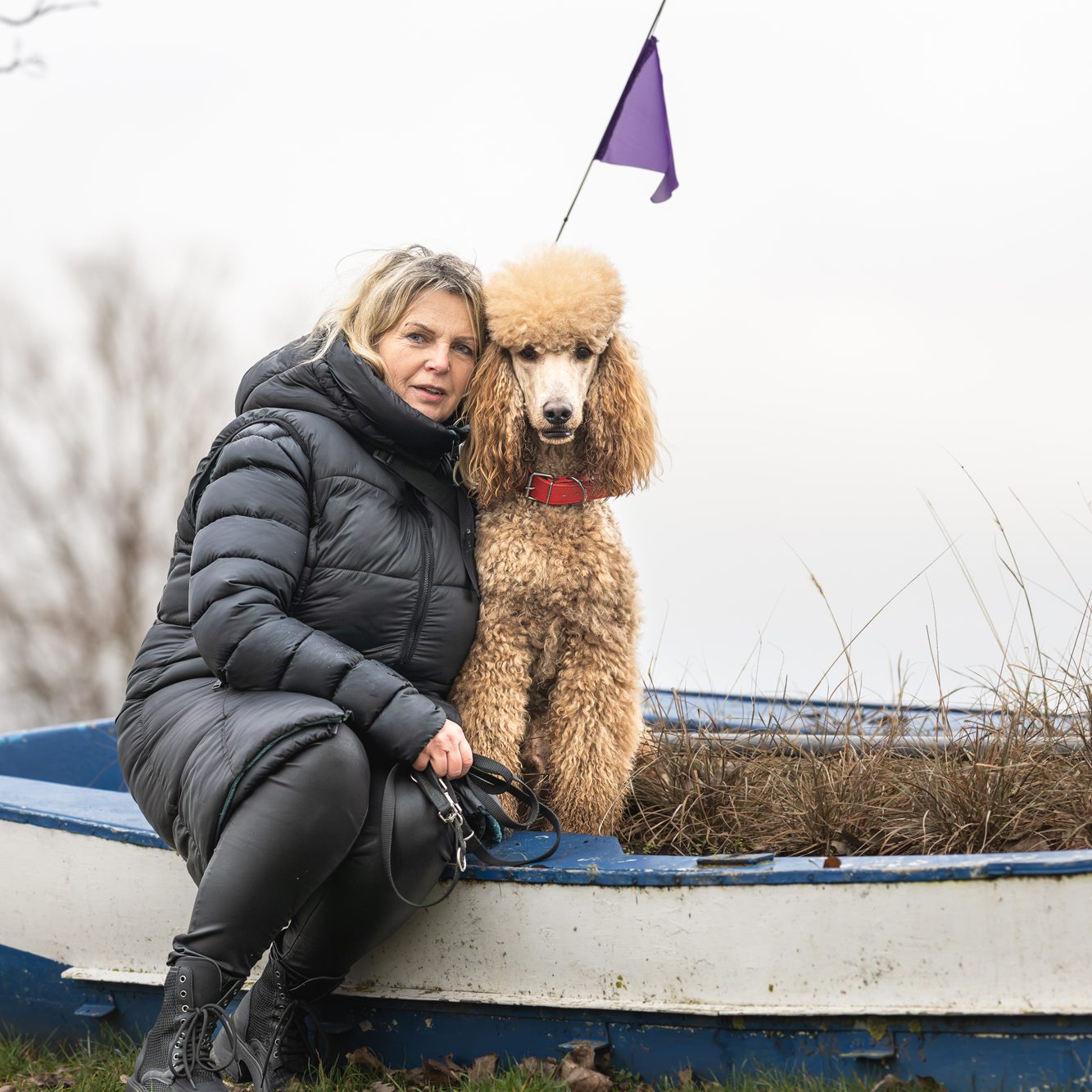 Ramona in schwarzer, warmer Jacke sitzt gemeinsam mit Großpudel Hadi am Rand eines alten Ruderbootes